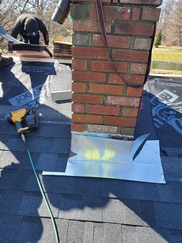 Roofers installing metal flashing around a brick chimney on a dark shingle roof.
