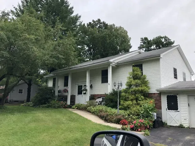 White two-story house with black trim, a walkway, and a small garage. Lush green lawn and trees.