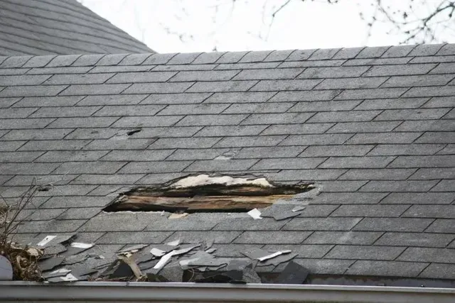 Damaged roof with a large hole exposing wood, broken shingles, and a gutter below.