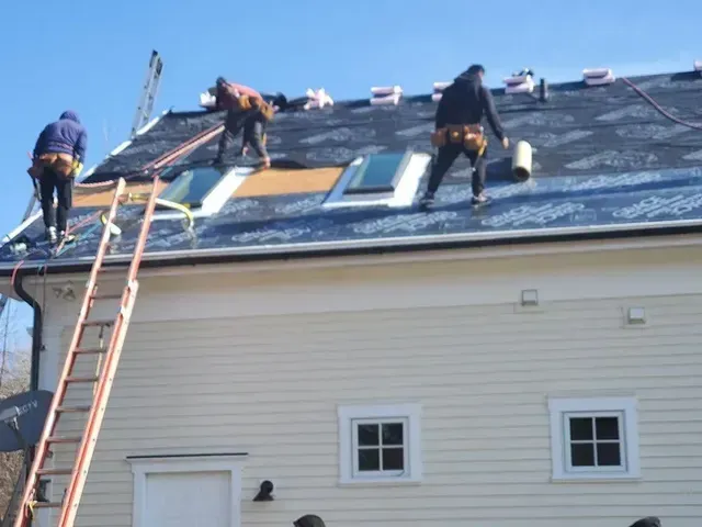 Three roofers on a roof, installing shingles. They are wearing safety harnesses and working near skylights.