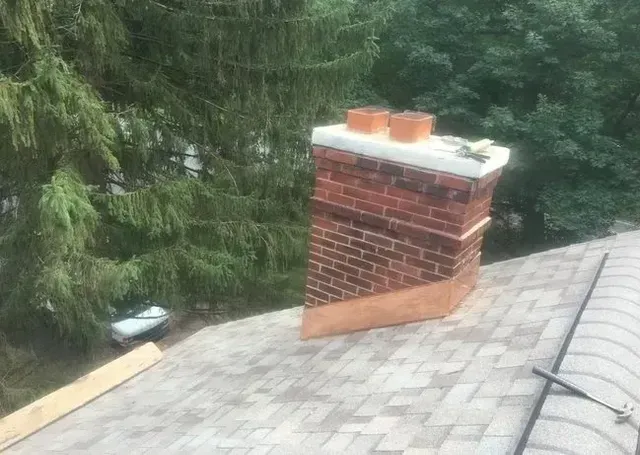Brick chimney on a shingled roof with copper flashing, two clay chimney pots, and surrounding trees.