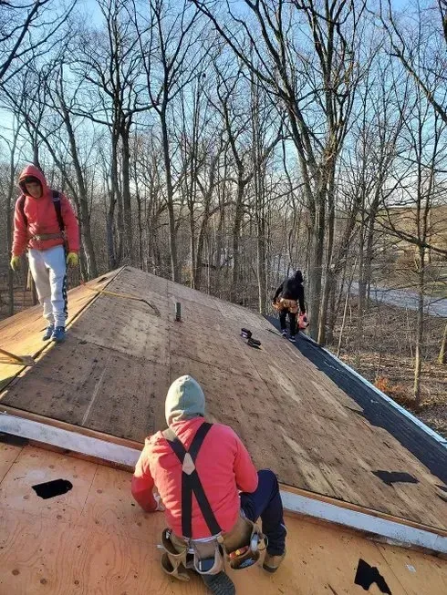Workers on a roof, installing shingles. One sits, two stand; red jackets, tan roof. Trees in background.