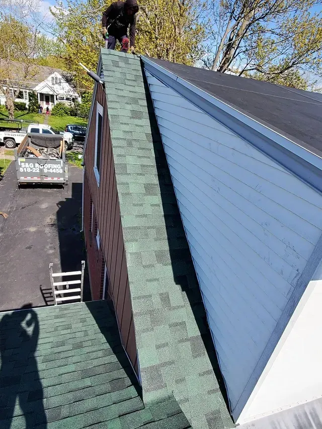 Person on a roof working on a chimney. Green shingles and light blue siding. Bright, sunny day.