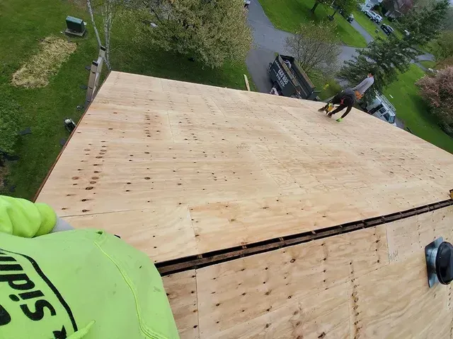 Roofer on a newly plywooded roof. A black truck and nearby houses are visible.