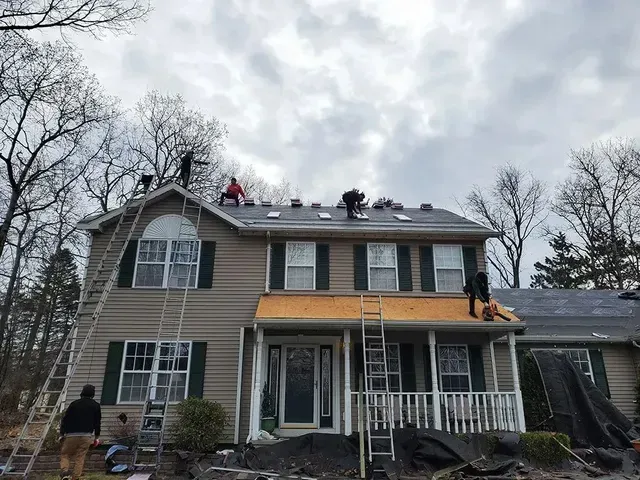 Workers replacing a roof on a two-story beige house with green shutters and overcast sky.