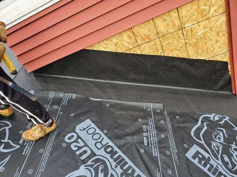 Person installing roofing material on a house; black underlayment with red siding visible.