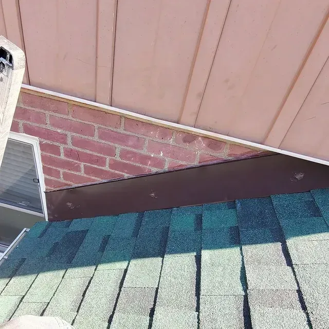 View of a roof with green shingles, brick wall, and brown flashing against pink siding.
