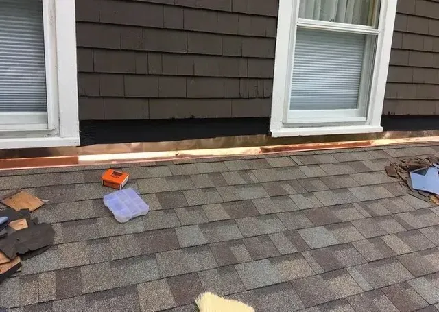 Roof with copper flashing around windows, brown siding, and asphalt shingles. A tape measure and tools are visible.