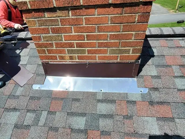 Chimney with brown and silver flashing on a shingled roof, person working on the left.