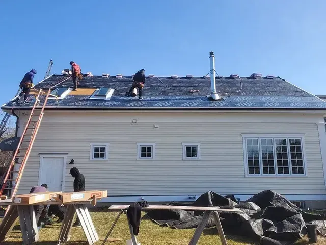 Roofers working on a house roof on a sunny day. They are using ladders, wearing work clothes, and there's a chimney.