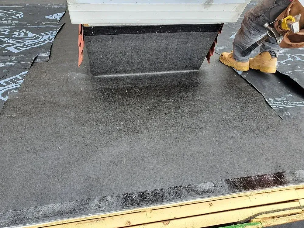 Roofer installing roofing membrane around a metal vent on a roof. A person's legs and work boots are visible.