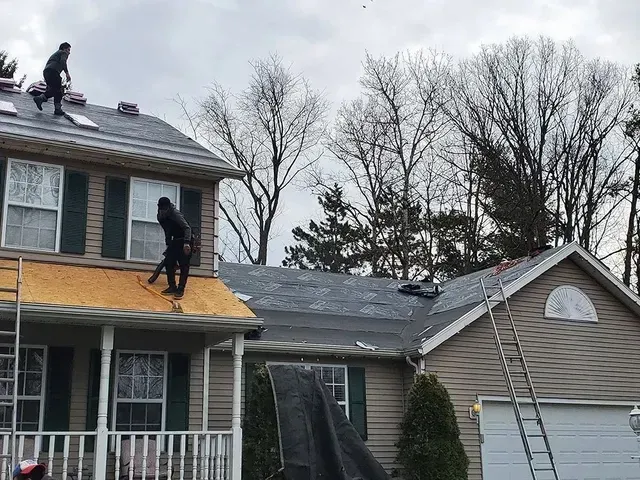 Roofers working on a residential home with ladders, tarps, and exposed roofing material. Overcast day.
