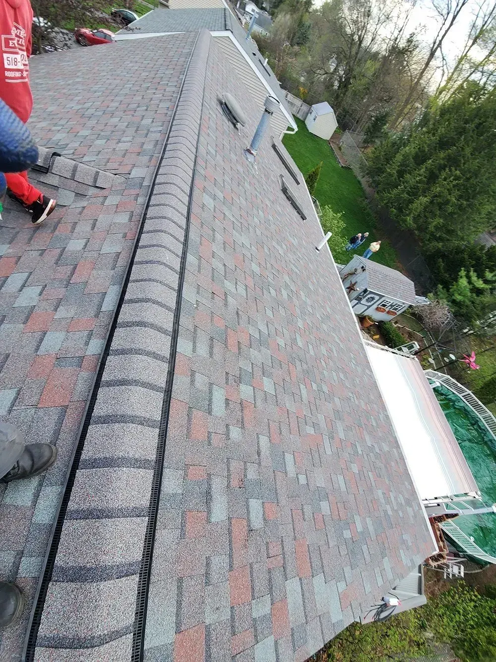 Rooftop with shingles in brown, gray, and red. Person standing on the roof, next to a curved edge. Green lawn visible.