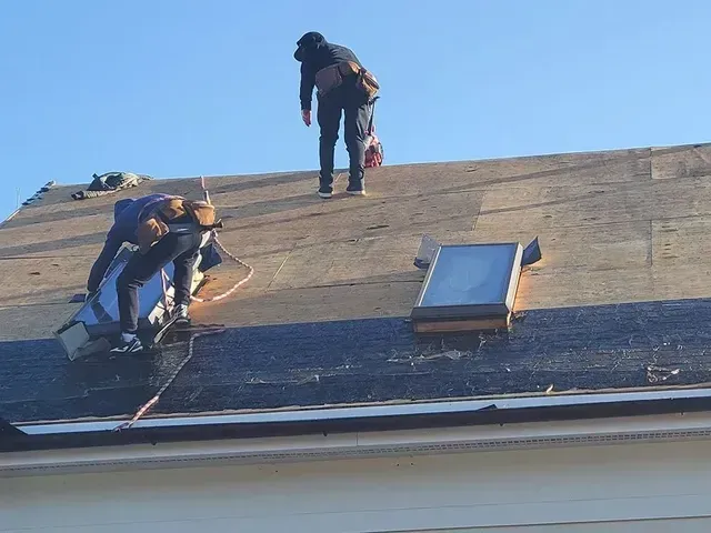 Construction workers on a roof, installing or repairing shingles. One worker is standing, two are working by a skylight.