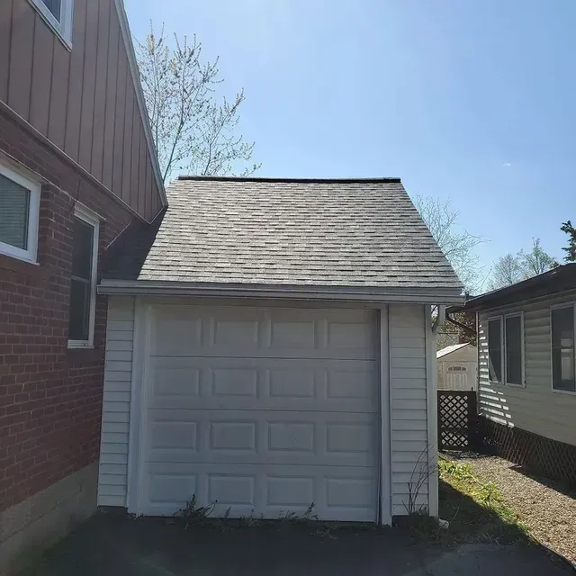White garage with gray roof, between brick house and other house on sunny day.