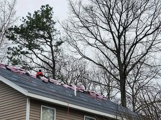 Roofer working on a gray shingled roof, pink insulation visible, trees in background, overcast day.