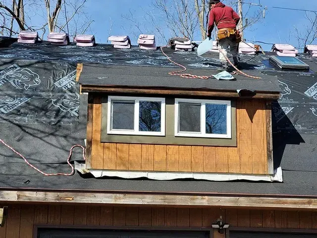 Roofer on a black roof, near a dormer with windows. Blue sky, tools visible.