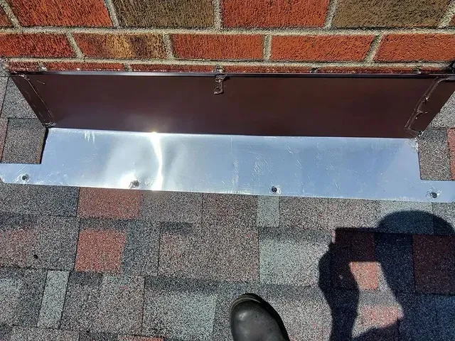 Metal flashing around a brown vent cover on a shingled roof, against a brick wall.