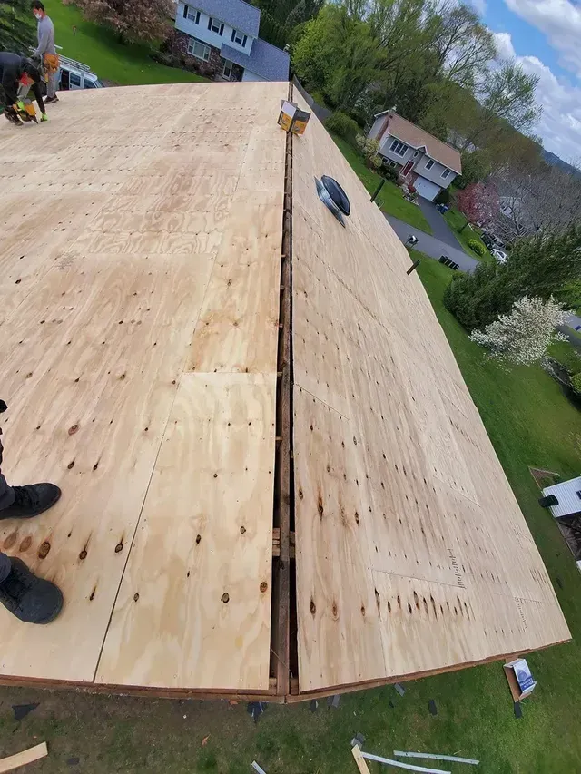 Newly installed plywood on a roof with a gap; a person is working nearby. Houses and trees in background.