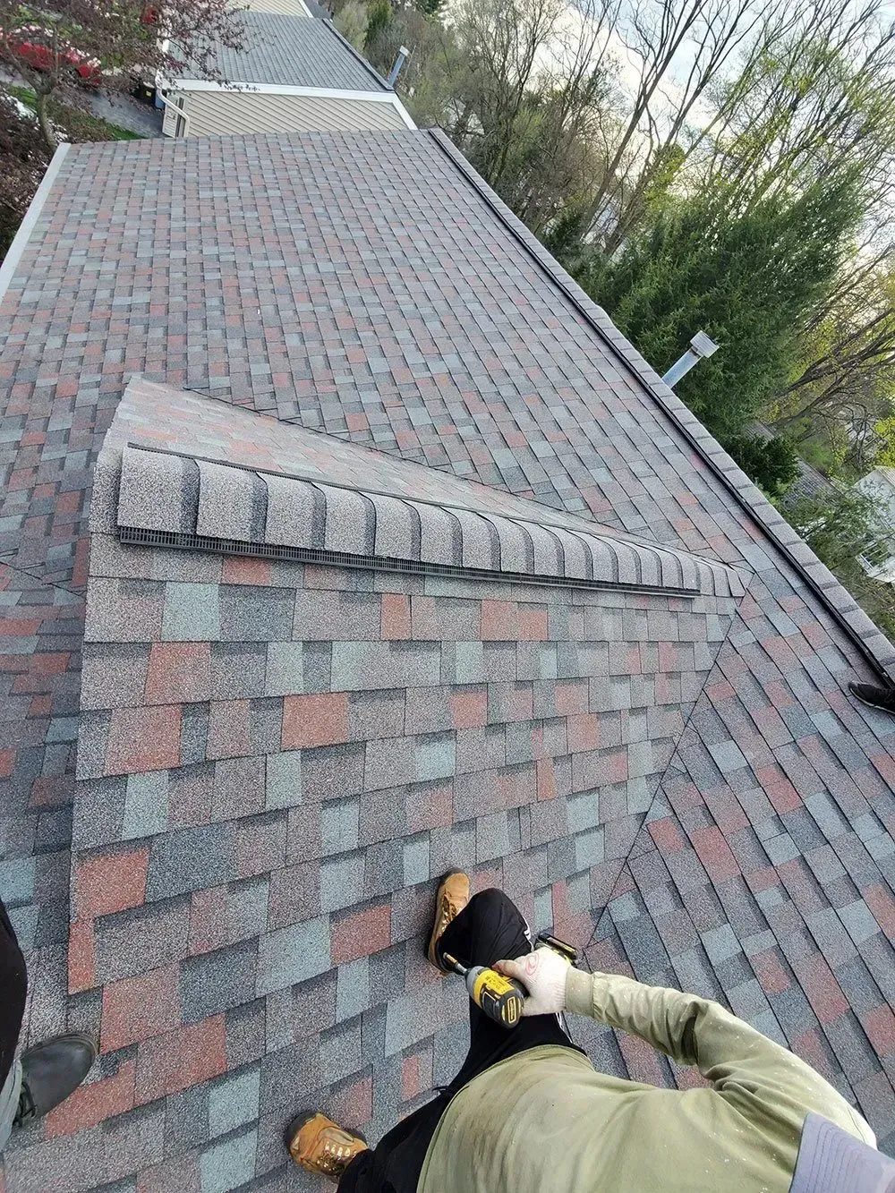 Person on roof measuring shingles near chimney. Brown, gray, and red shingles. Green trees in the background.