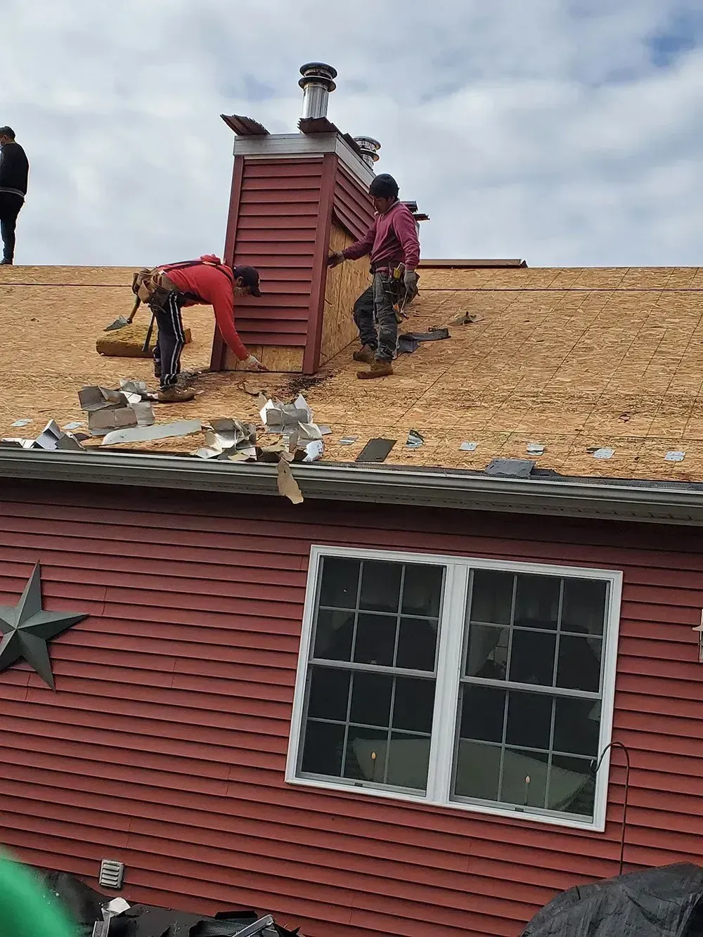 Two roofers on a rooftop near a chimney, working on roof shingles. The house is red.