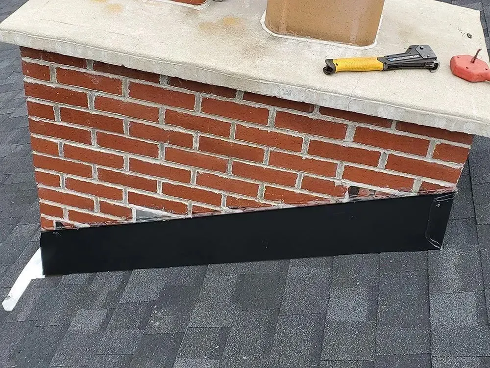 Brick chimney with black flashing against gray roof. Tools rest on the chimney's top surface.