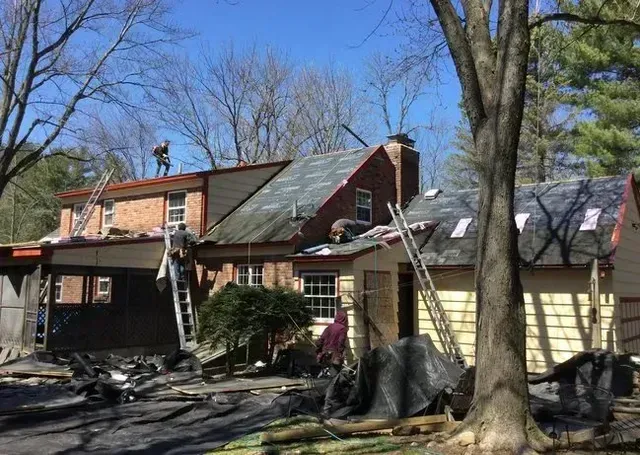 Construction workers removing and replacing roof shingles on a two-story brick home, ladders present.