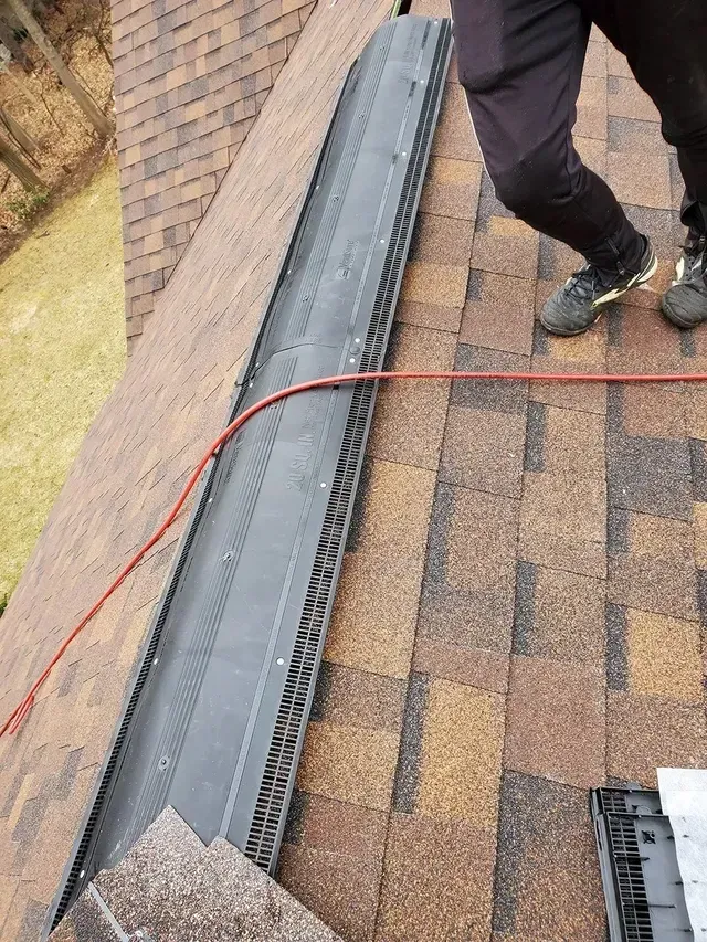 Roofer installing a ridge vent on a brown shingle roof. Gray vent, red cord, person's legs visible.