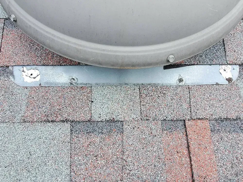 Metal bracket securing a dome-shaped object to a shingled roof.