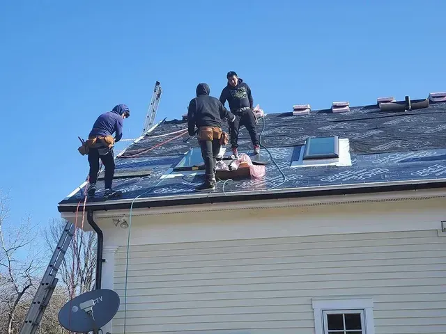 Three roofers working on a rooftop, securing materials; clear sky, sunny day.