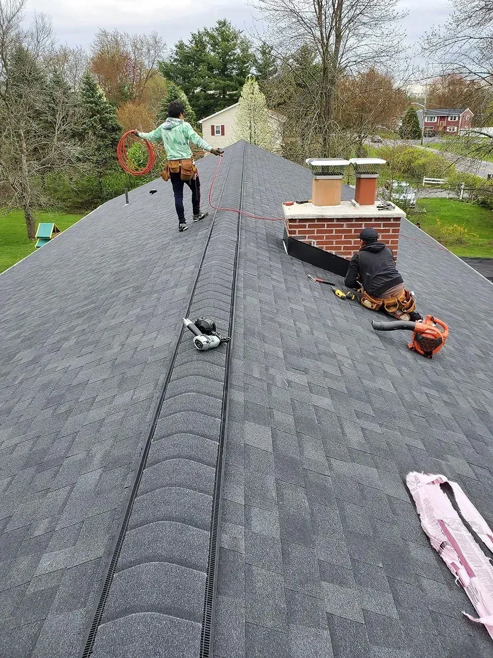 Two people working on a dark gray shingled roof, one near a chimney, one walking. Autumn trees in the background.