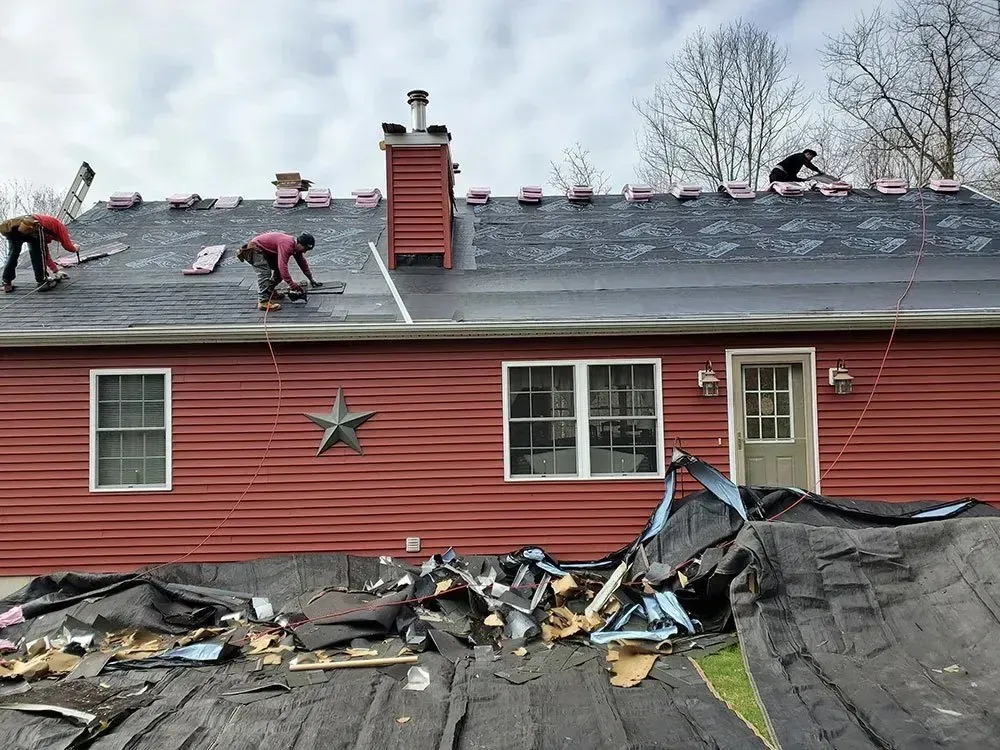 Roofers working on a red house roof, removing old shingles. Black tarp covers ground.