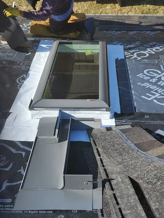 Roofer installing flashing around a skylight on a shingle roof; gray metal flashing, black underlayment, and shingles.
