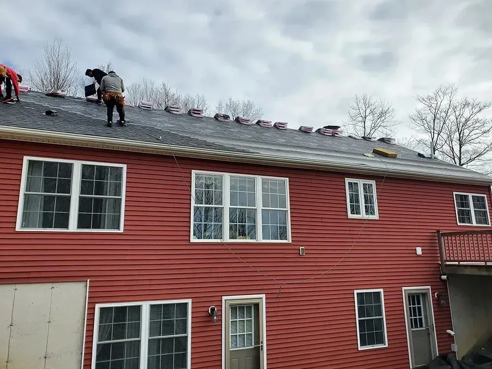 Roofers installing shingles on a red-sided house with multiple windows on an overcast day.