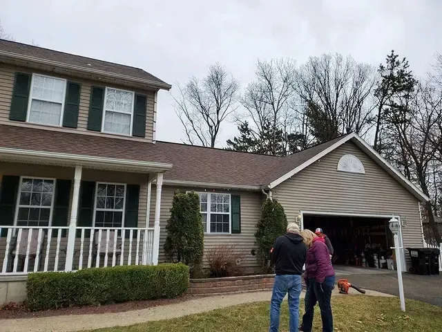 Two-story house with two people in front; one with a leaf blower. Gray sky, green lawn and shrubs.
