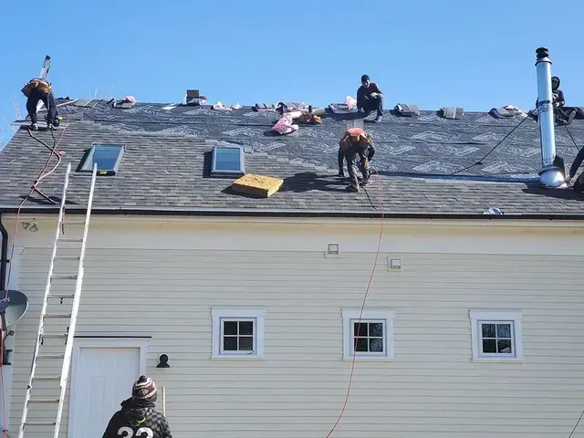 Roofers working on a house roof under a blue sky, some on ladders, removing old shingles.