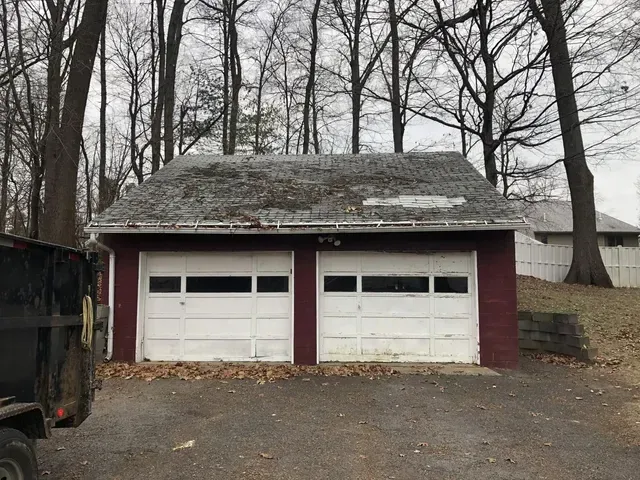 Two-car garage with white doors, red trim, and a deteriorating gray roof, set against a backdrop of trees.