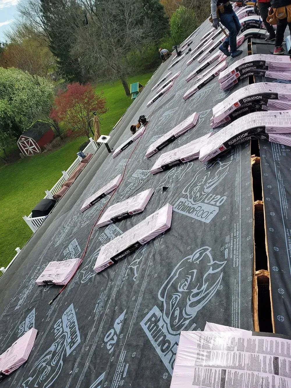 Roofers installing shingles on a roof, with pink insulation visible. Green grass and trees in background.