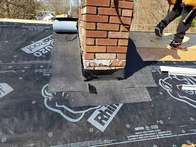 Roofer installing flashing around a brick chimney on a roof. Black underlayment visible.