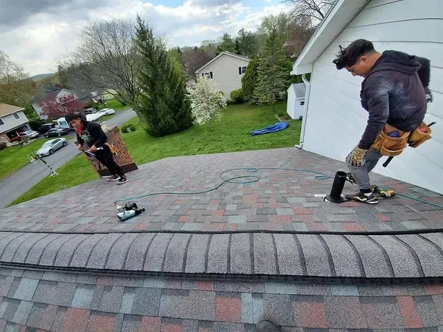 Two people installing shingles on a roof; one holds a bundle, the other works with tools. Overcast day.
