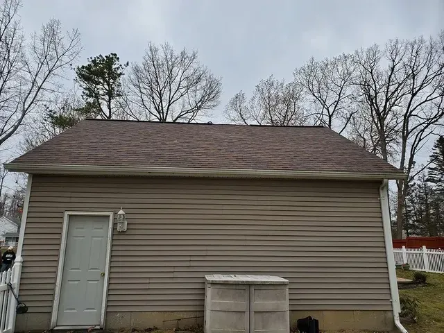 Tan-sided building with brown roof and white door, surrounded by trees under a cloudy sky.