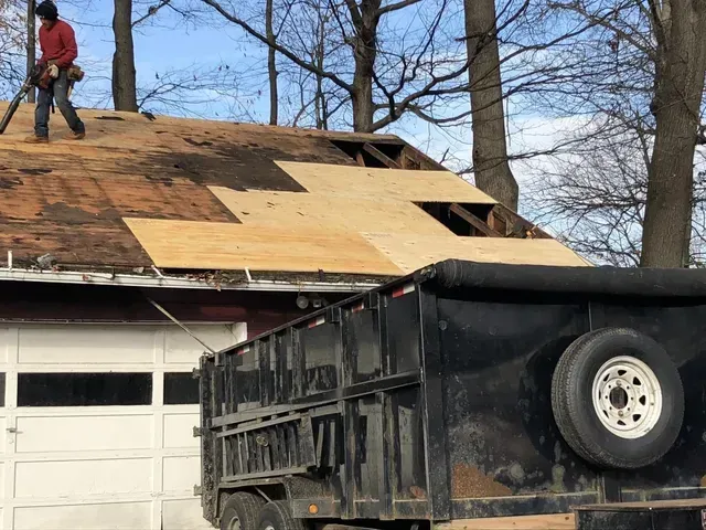 A person on a roof replacing plywood next to a black trailer, in front of a white garage.