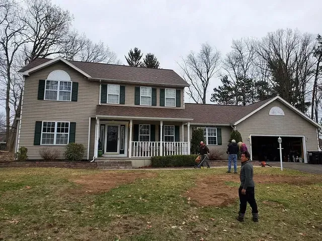 Two-story beige house with green shutters, a porch, and attached garage. People are in the yard, working.