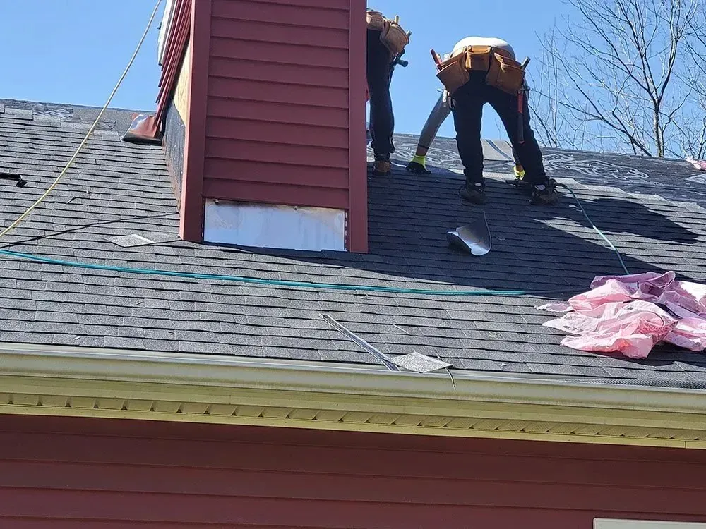 Two workers on a roof repair a red chimney, visible sky, shingles, and safety lines.