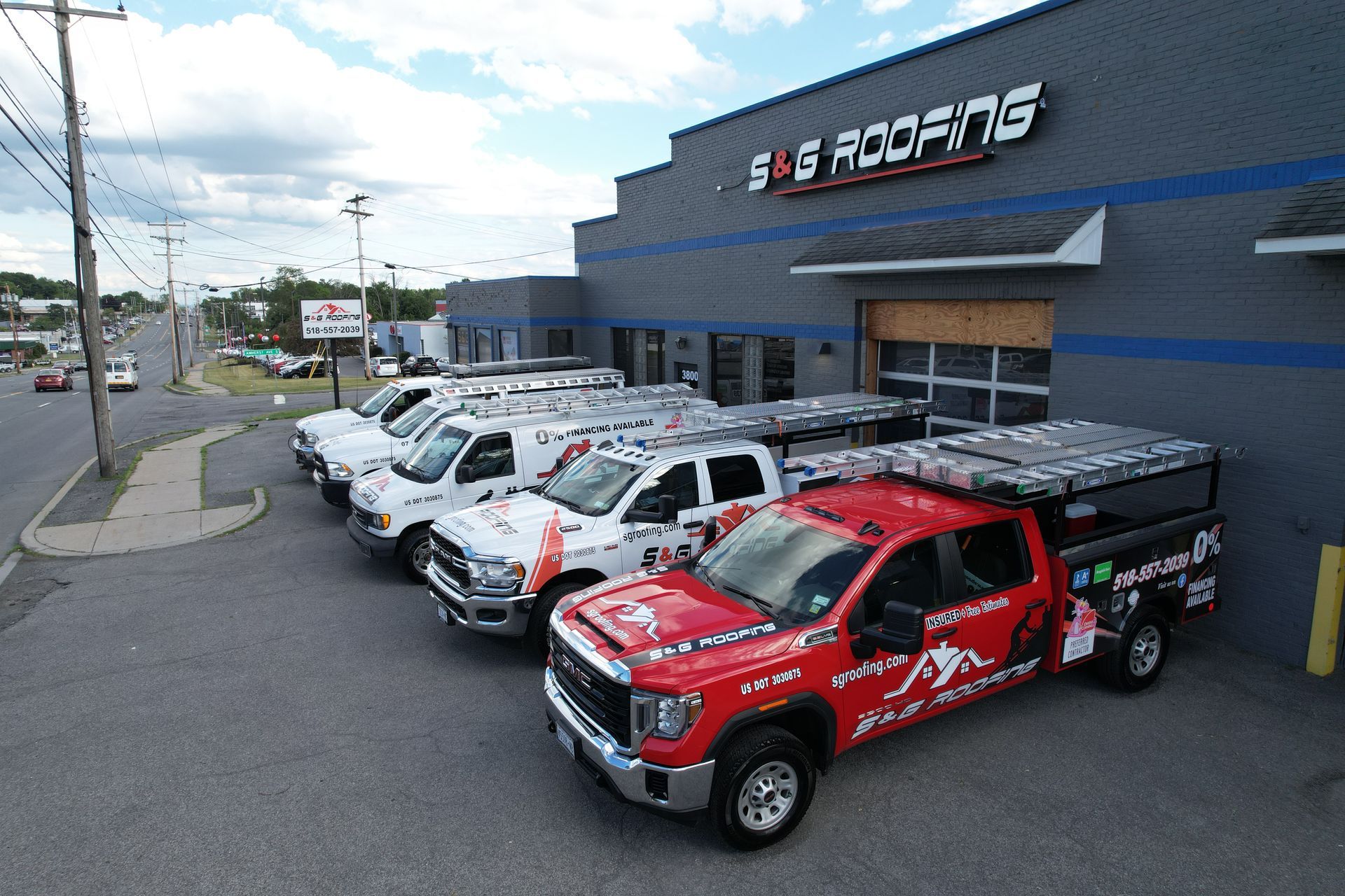 Several white and red work trucks parked in front of a gray roofing company building under a cloudy sky.