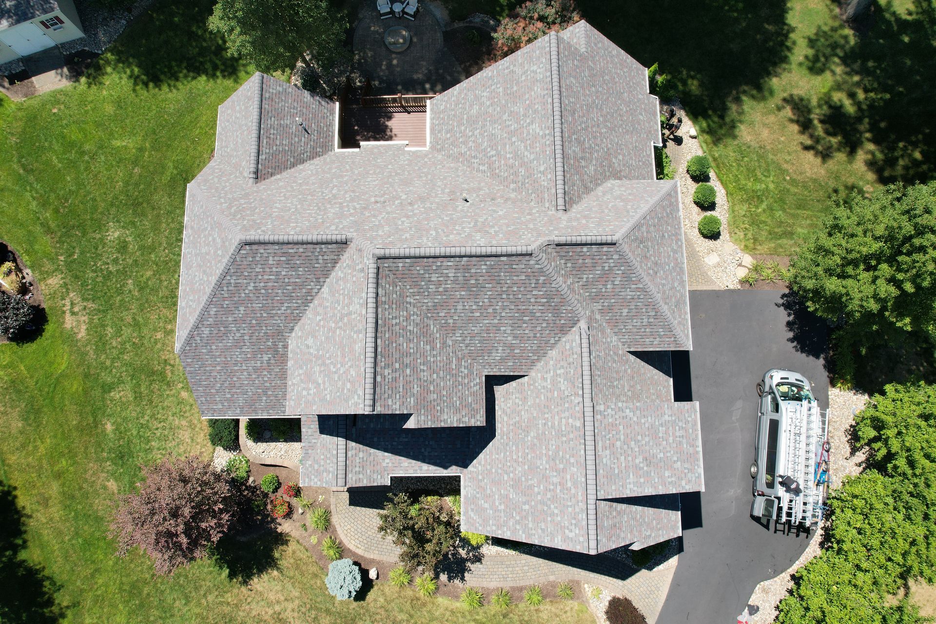 Overhead view of a house with a gray roof, surrounded by green grass and trees. A vehicle is in the driveway.