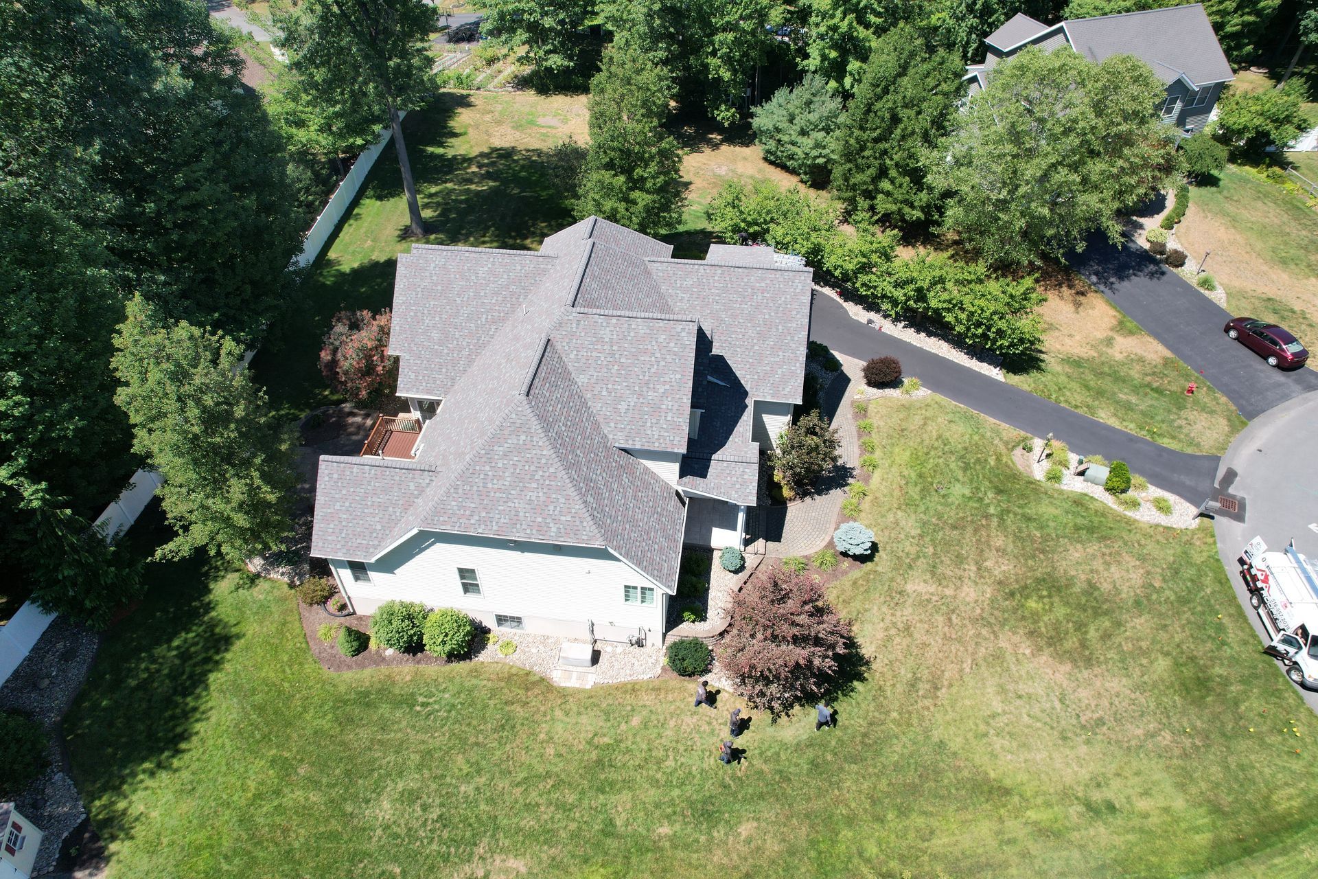 Aerial view of a gray-roofed house with a long driveway surrounded by green trees and grass on a sunny day.