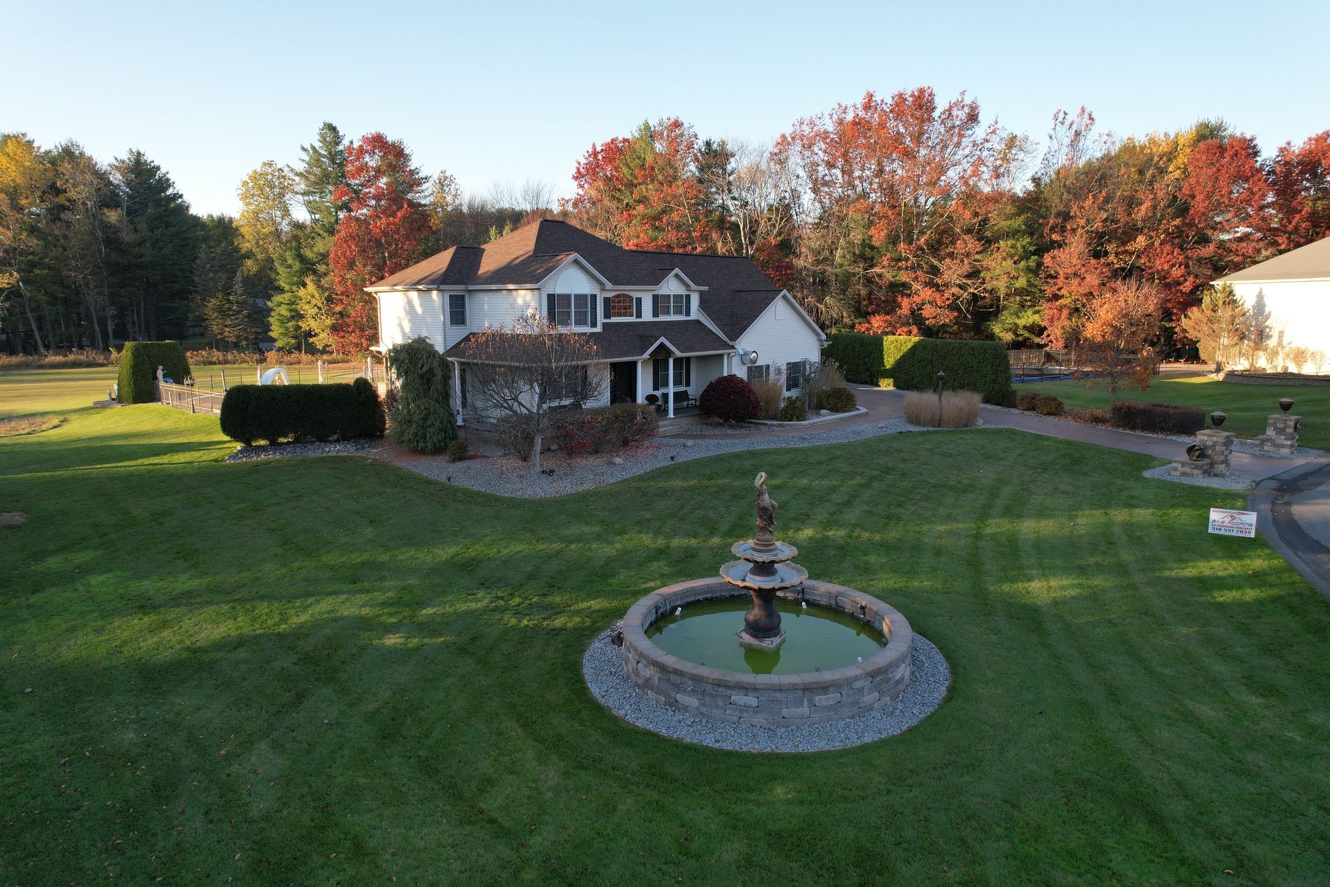 Two-story white house with fall foliage, fountain in front lawn, sunny day.