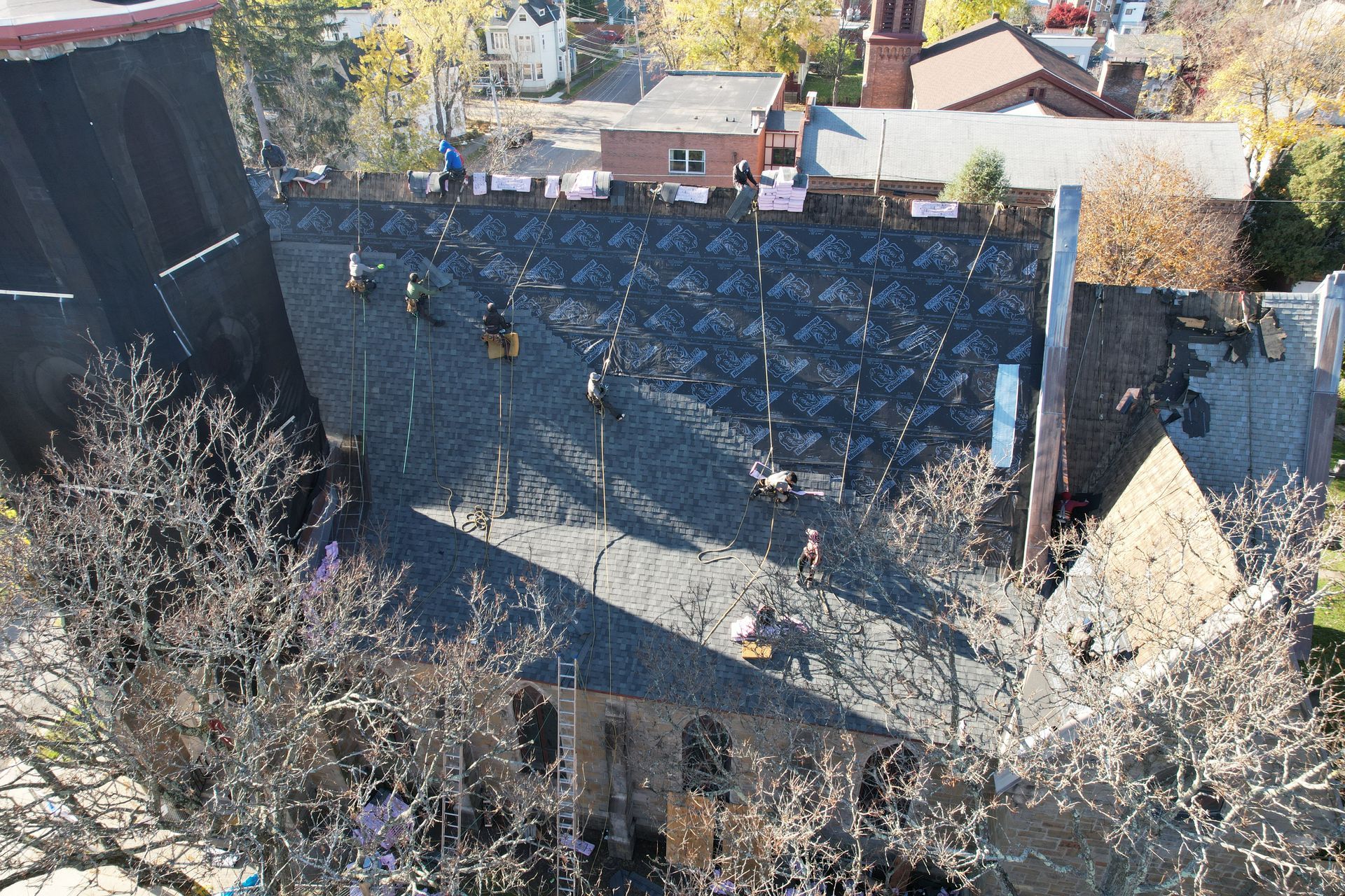 Roofers on a dark, angled roof; surrounding buildings and trees in view.