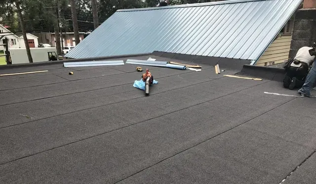 Workers installing roofing material on a flat roof next to a metal roof section.